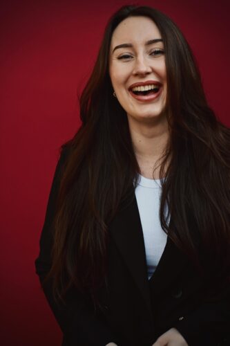 Kim is een jonge vrouw met donkerbruin haar die met open mond lacht. Ze heeft een zwart jasje aan met een wit shirt. Kim staat voor een donkerrode achtergrond.